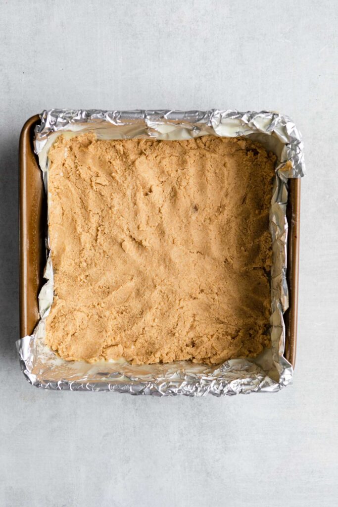 cookie dough pressed into a square foil lined baking pan on a grey surface
