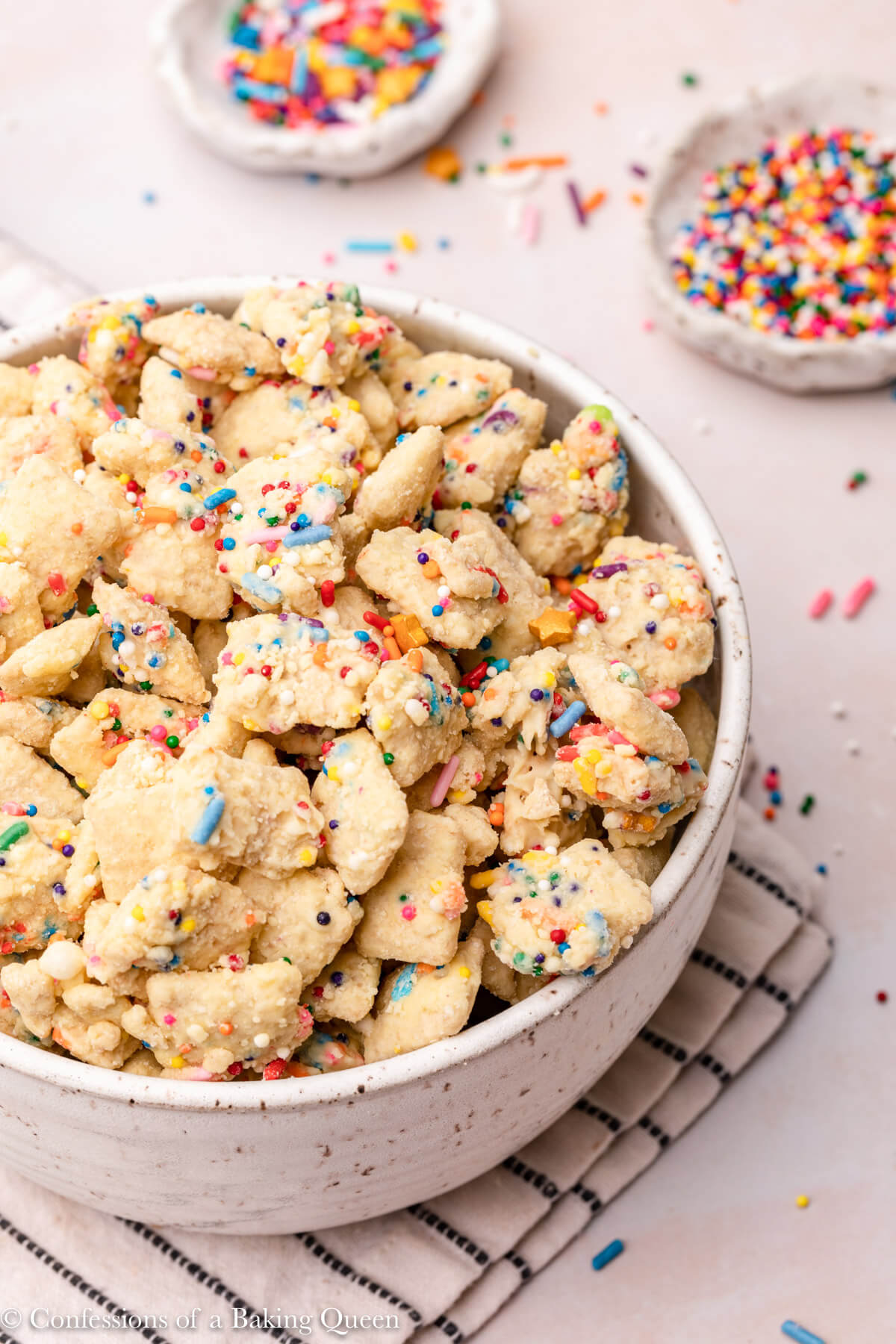 cake batter muddy buddies in a large white bowl on top of a white and blue napkin on a light pink surface