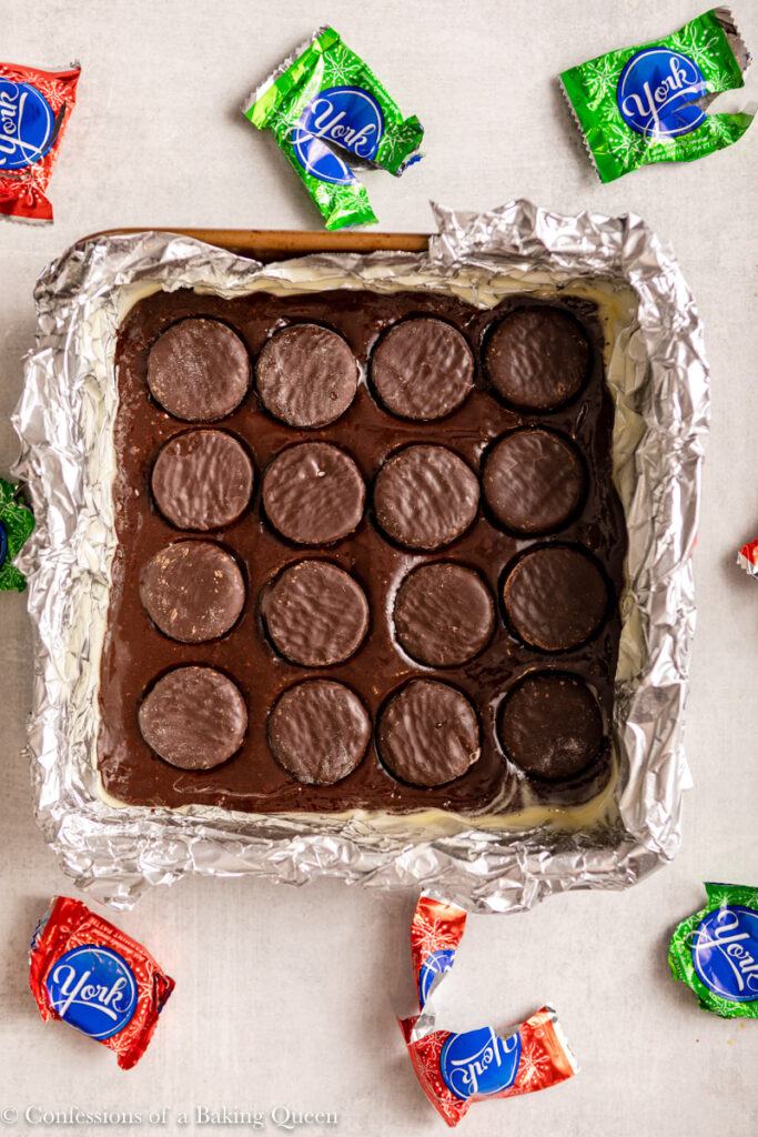 brownie batter poured into pan and peppermint patty's added on top in a foil lined pan on a light grey surface with peppermint patty wrappers
