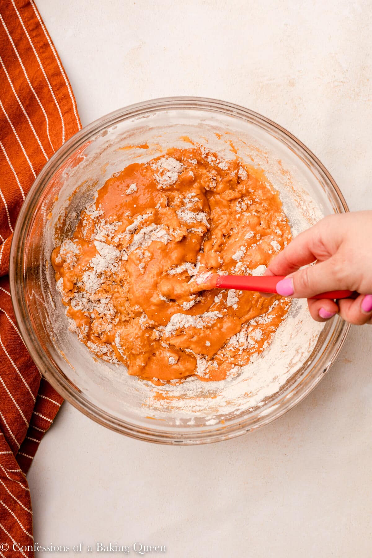 hand holding a spatula gently folding dry ingredients into wet ingredients on a light surface with an orange linen