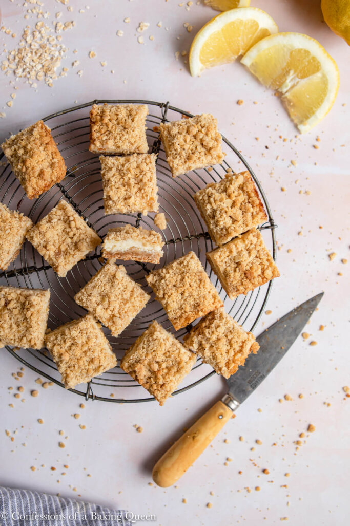 lemon crumb bars on a wire rack on a light pink surface with lemon slices, a knife, and a blue linen