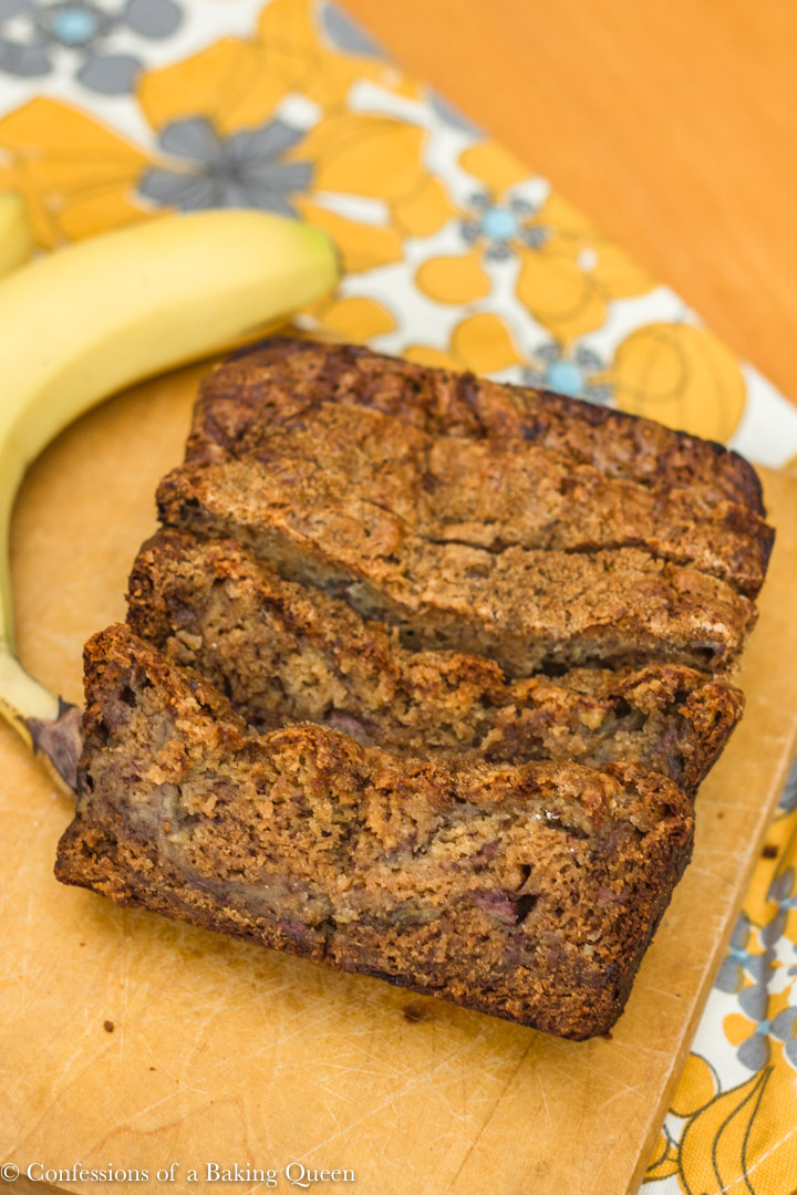 sliced pudding banana bread loaf on a wood board with a banana next to it all sitting on a grey white and yellow tea towel