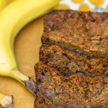 cut slices of pudding banana bread on a wood board with a banana next to it all sitting on a yellow grey and white tea towel