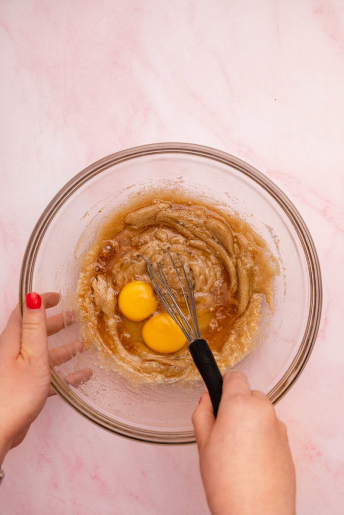 hand holding a whisk mixing eggs into butter sugar mixture in a glass bowl 