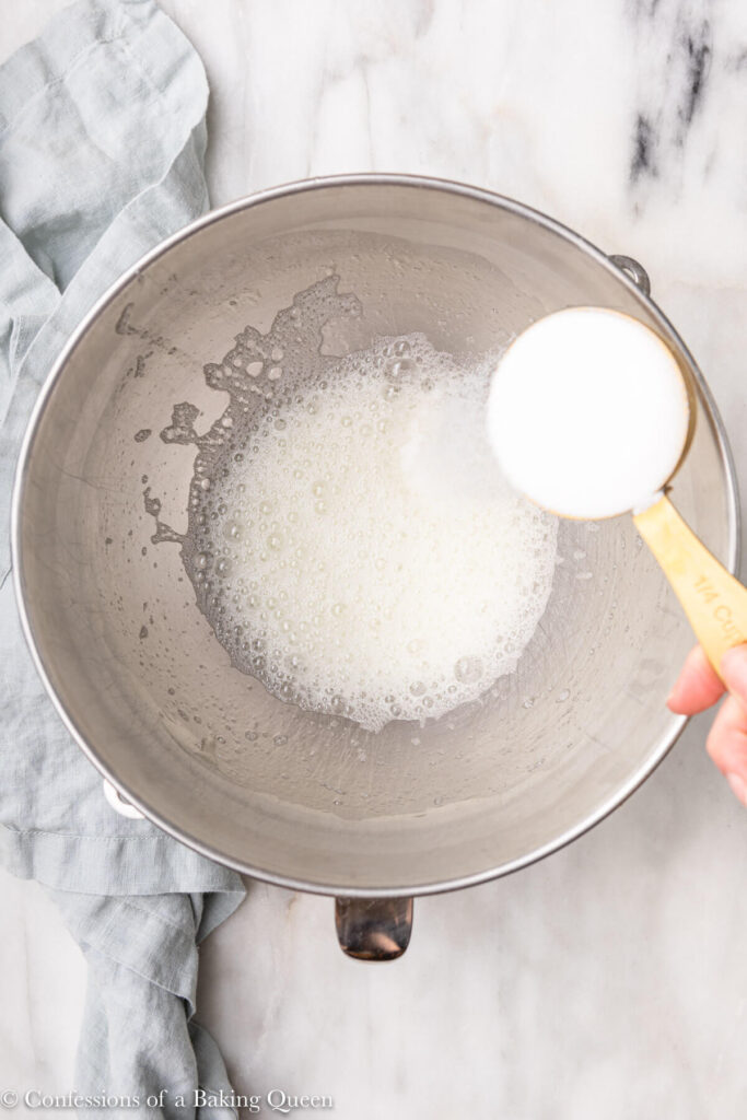 sugar slowly added to foamy egg whites in a metal mixing bowl on a white marble surface with a light green linen