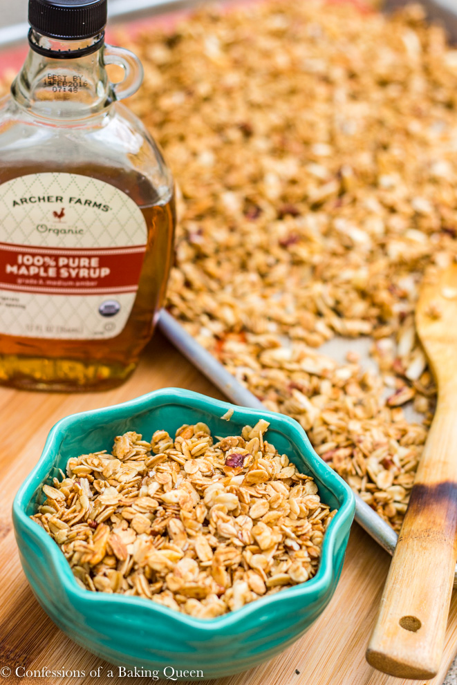 homemade maple granola recipe on a baking sheet and in a turquoise bowl with a bottle of maple syrup all on a wood board