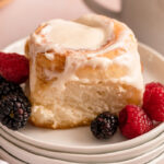 cinnamon roll on a stack of white plates with berries on a light pink surface with a cup of coffee and linen