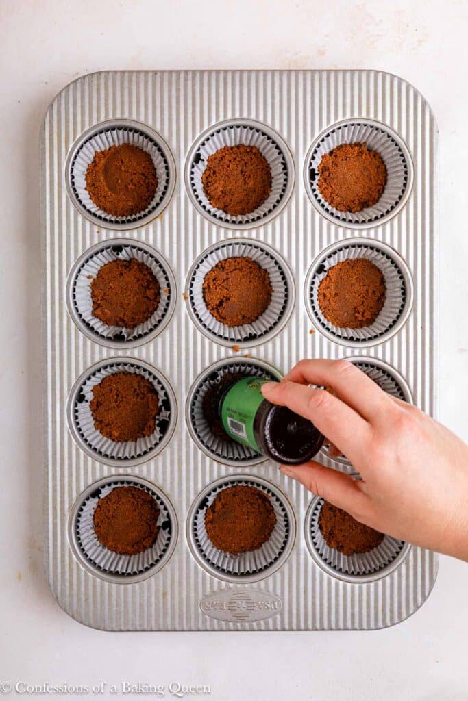 spice jar pressing cookie crumbs into cupcake liners in a muffin tin on a light surface.
