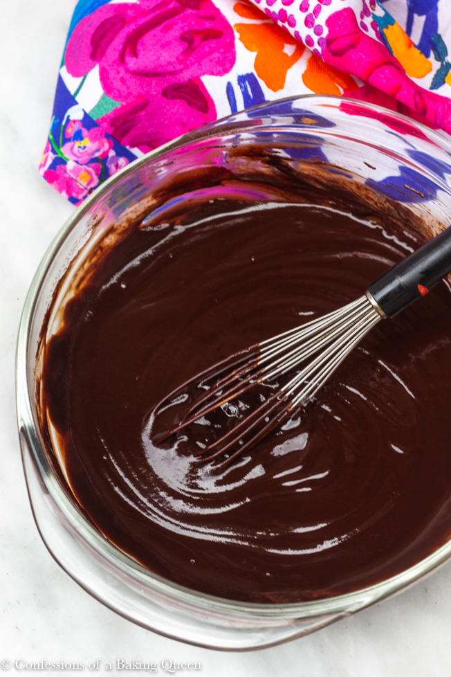 chocolate ganache in a glass bowl with whisk on a white surface with a multi-colored linen