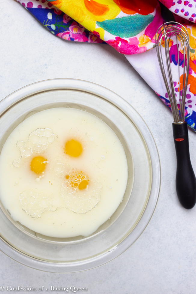 eggs with the rest of the wet ingredients in a bowl with a small whisk next to bowl with a multi-colored linen and whisk on a white background
