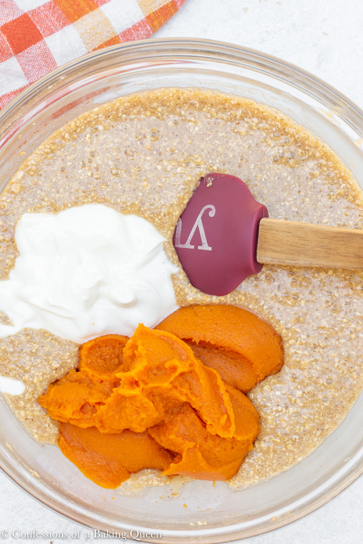 pumpkin and yogurt added to oat flour pumpkin bread batter in a glass bowl on a white background with an orange, yellow, and white linen