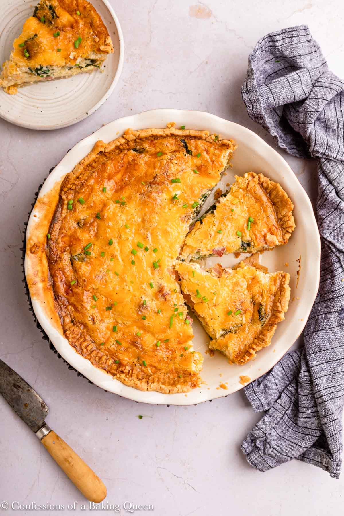 cut up bacon cheddar quiche in a white pie dish with a wooden knife on a light surface with a blue linen.