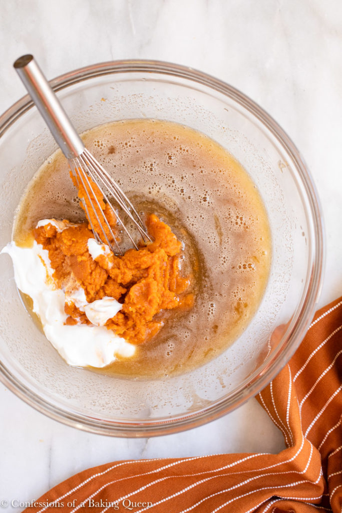 pumpkin puree and Greek yogurt added to wet ingredients in a large glass bowl on a white surface with an orange and white linen