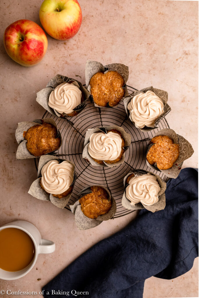 apple cupcakes with maple frosting on a wire rack on a light brown surface with a navy linen and apples and cup of coffee