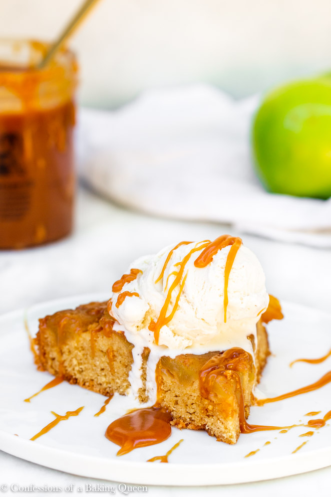 slice of upside down caramel apple cake on a white plate with ice cream and caramel drizzle on a white surface with a caramel cup and green apple in the background