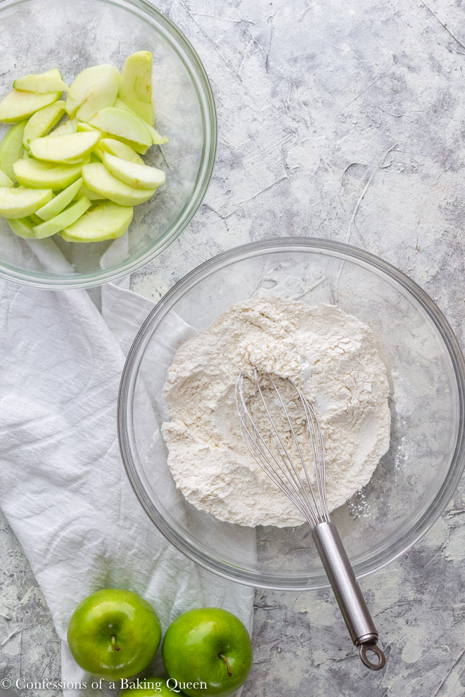 dry ingredients whisked together in a glass bowl and another bowl of sliced apple slices on a grey surface with a white linen and two green apples