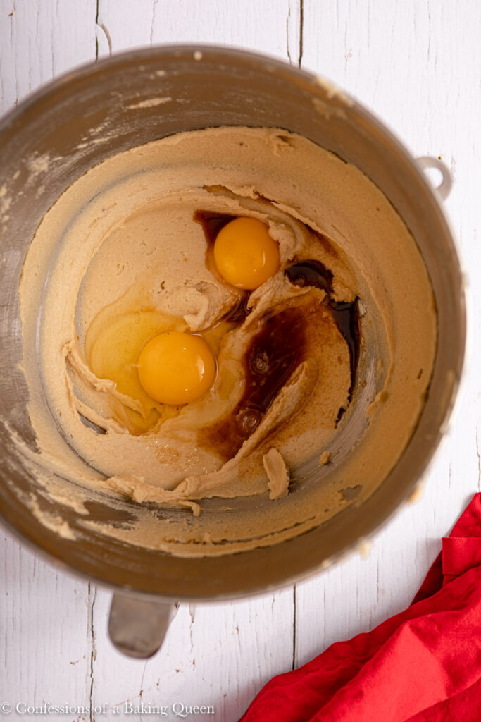 eggs and vanilla added to creamed butter mixture in a metal mixing bowl on a white wood surface with a red linen