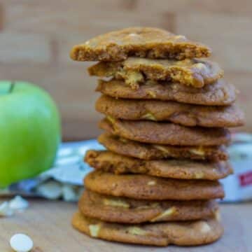 Salted Caramel Apple White Chocolate Cookies stacked on top of each other on a wood board next to an apple