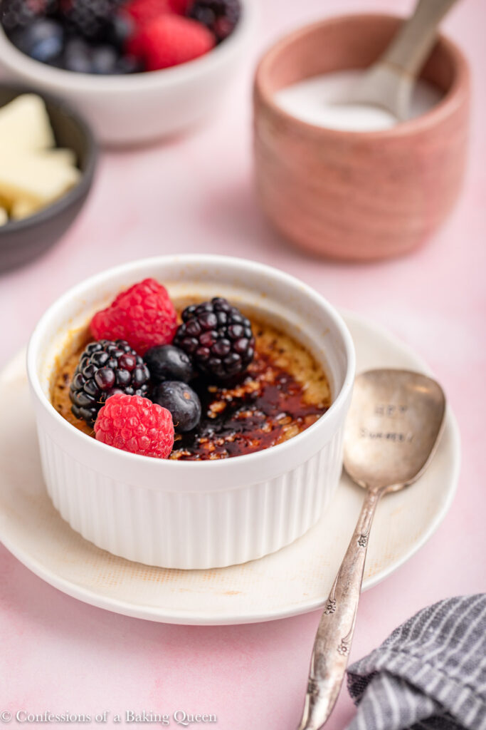 white chocolate creme brulee on a plate with a spoon next to a cup of sugar, bowl of berries and cup of white chocolate on a light pink surface