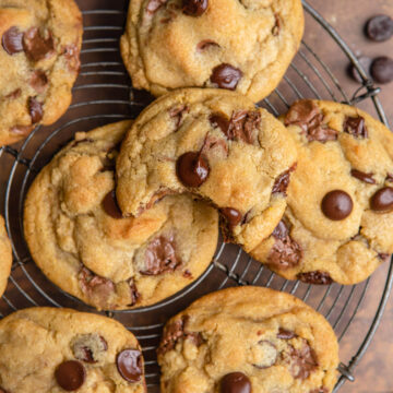 close up of easy chocolate chip cookies on a wire rack on a brown and orange distressed surface next to a blue linen and a dark bowl with chocolate chips spilled out