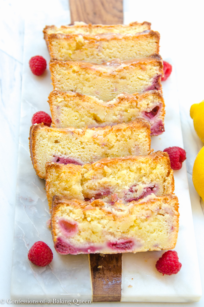 raspberry lemon loaf cake sliced served on a white and wood board with raspberries on a white surface with two lemons off to the side