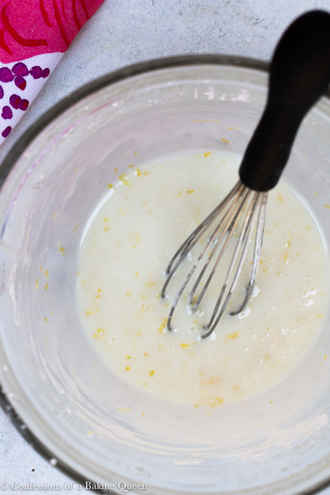 lemon glaze being whisked together in a clear bowl for raspberry lemon loaf cake on a light grey/ white background with a multi-colored linen