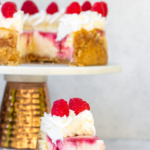 raspberry swirl lemon cheesecake slice on a white plate with the full cheesecake on a stand in the background on a white surface