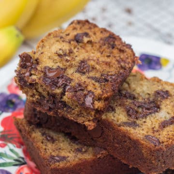 close up of baked slice of Chocolate Chip Banana Bread on a colorful plate with bananas and more banana bread in the background