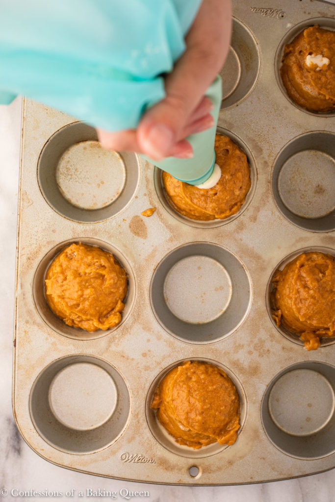 hand piping cream cheese into pumpkin muffin batter in a cupcake tin