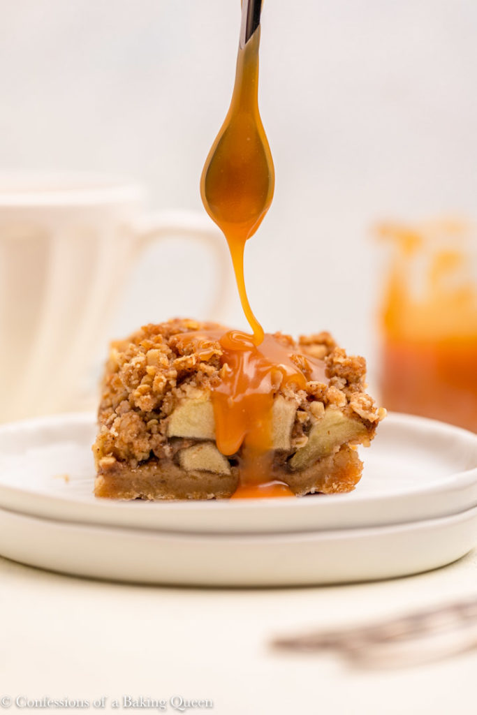 spoon drizzle salted caramel on an apple pie bar on two white plates on a white background with a cup of tea and more caramel in a clear jar