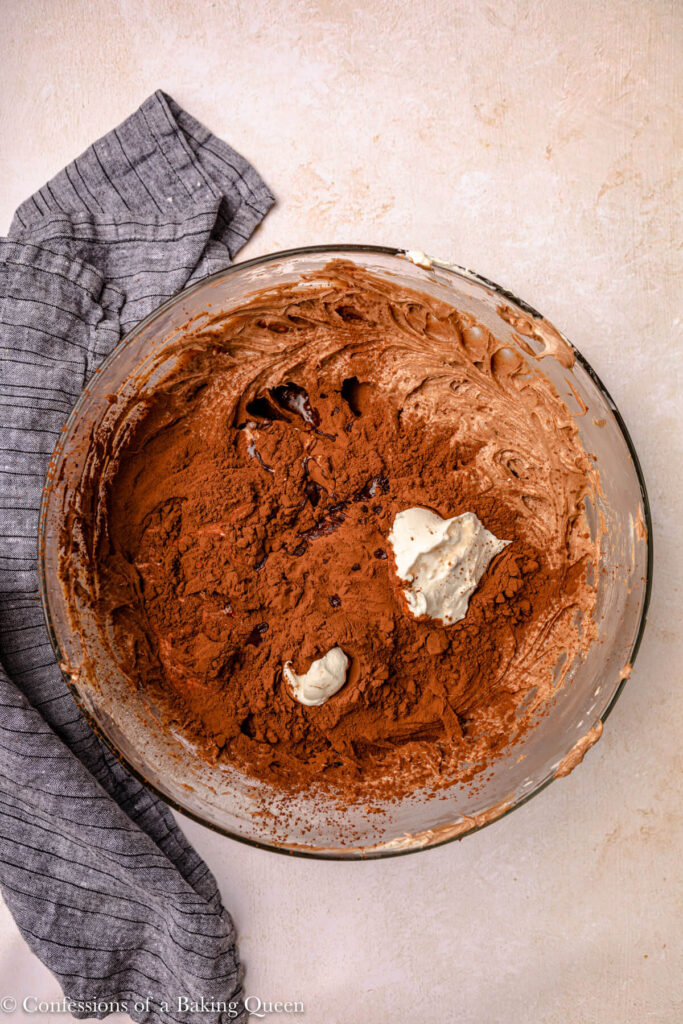 cocoa powder, heavy cream, and sour cream added to cheesecake batter in a glass bowl on an light pink surface with a blue linen