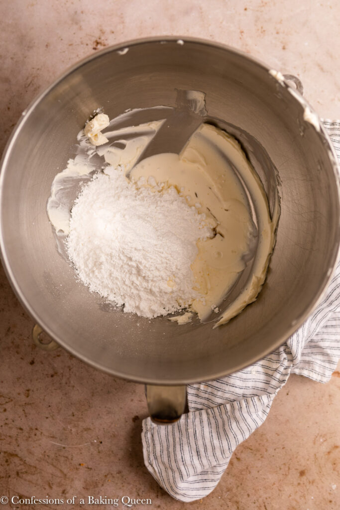 powdered sugar, heavy cream, and vanilla added to cream cheese in a metal mixing bowl next to a white linen on a light brown surface