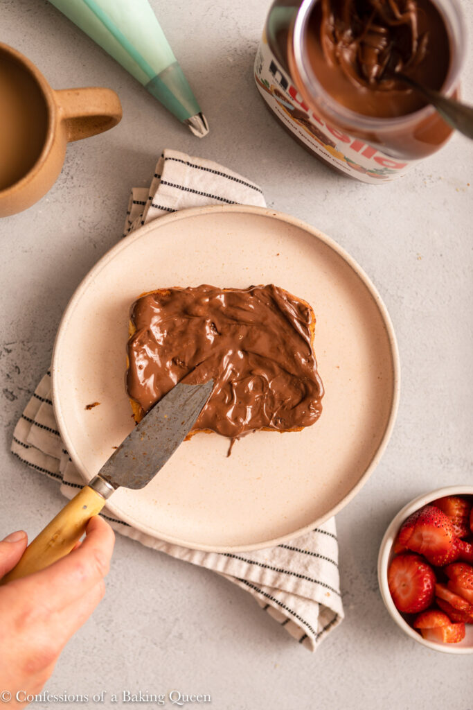 knife spreading nutella on to a slice of french toast on a light grey surface next to a jar of nutella, cup of coffee, and bowl of strawberries