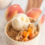 brown butter peach crumble in a cream ceramic dish with a fork taking a bite on a wood and marble board