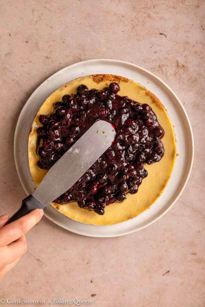 flat spatula spreading blueberry sauce on top of cheesecake on a white plate on a light brown surface