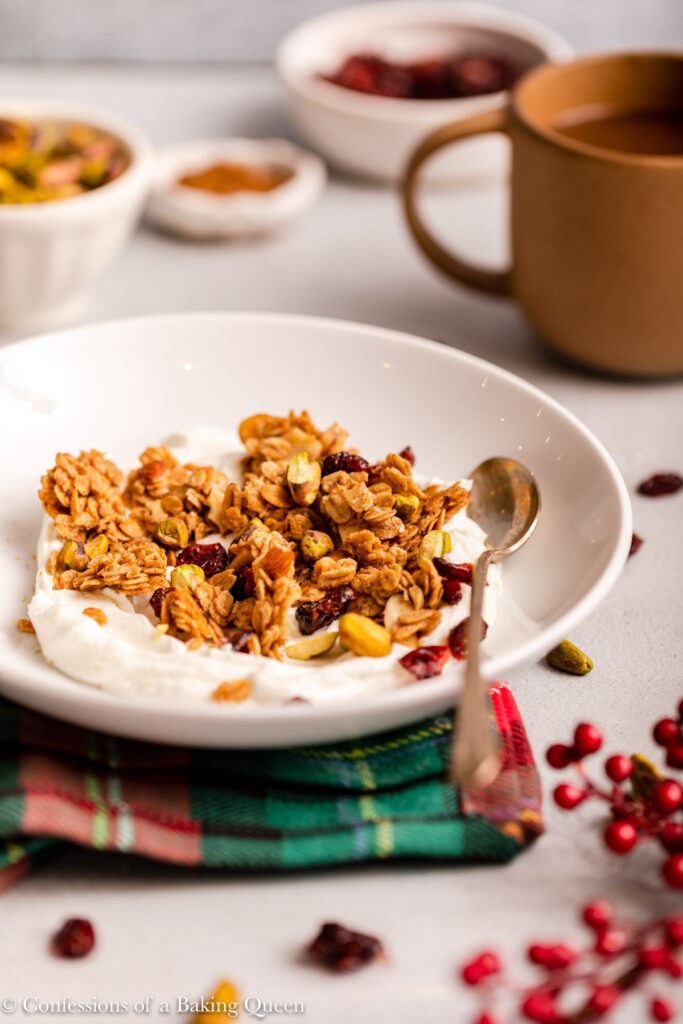 granola and yogurt in a bowl on a green and red checkered linen next to ingredients and coffee cup on a grey background