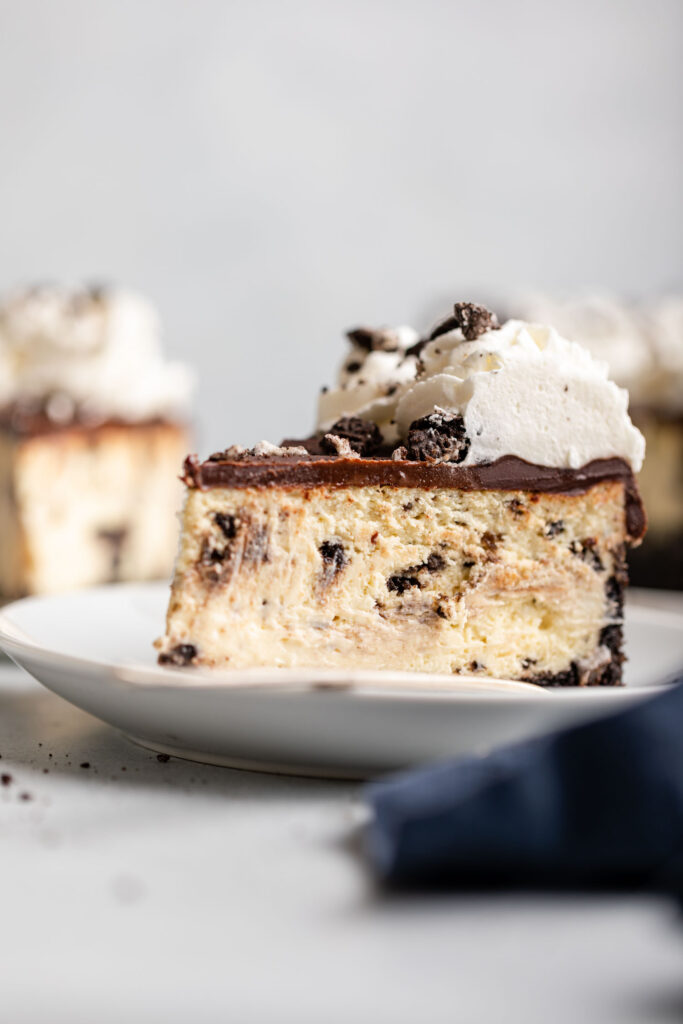 a slice of oreo cheesecake on a white plate with more cheesecake in the background sitting on a grey surface with a navy blue linen