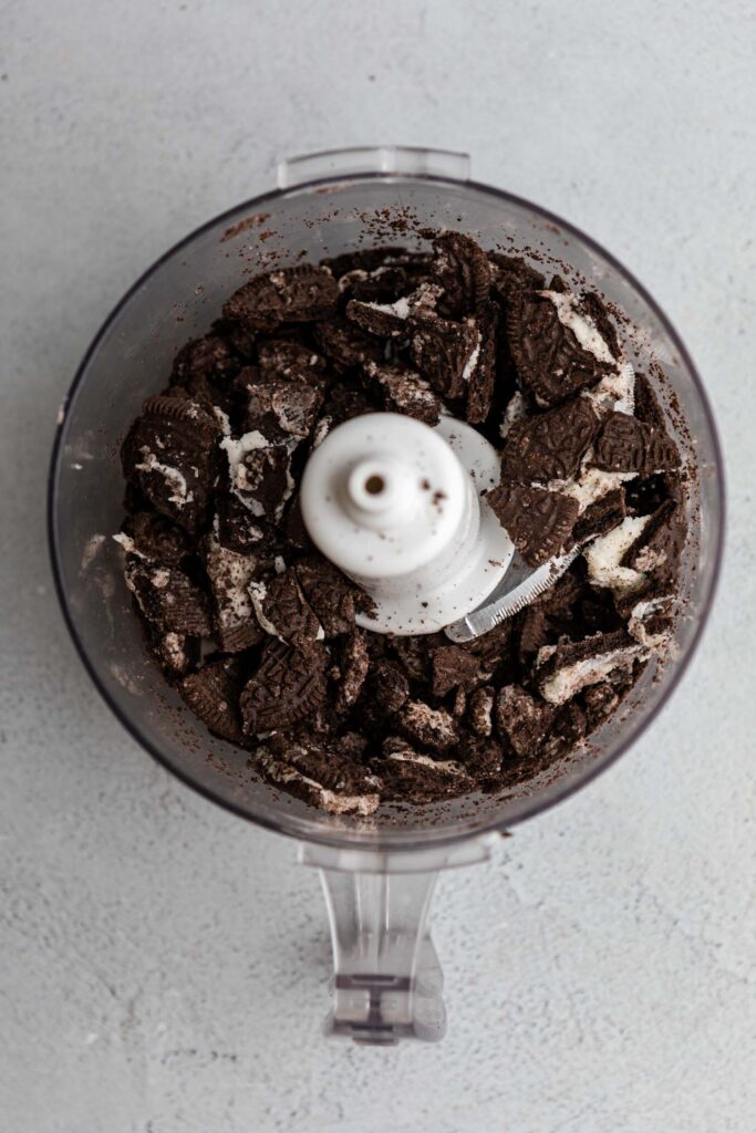 crushed oreos in a food processor then added to a cheesecake batter in a metal mixing bowl on a grey surface (