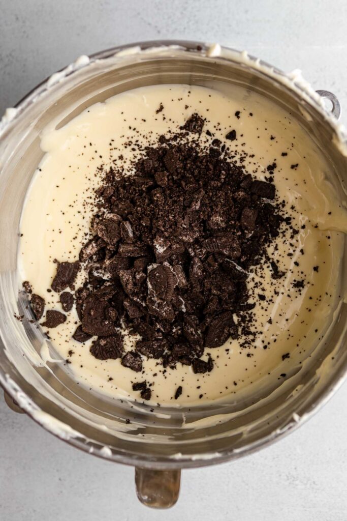 crushed oreos in a food processor then added to a cheesecake batter in a metal mixing bowl on a grey surface