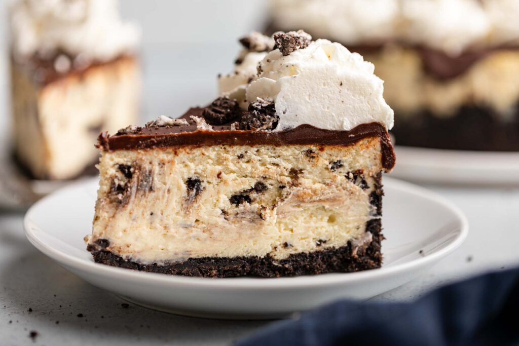 slice of oreo cheesecake on a white plate with more cheesecake in the background sitting on a grey surface with a navy blue linen
