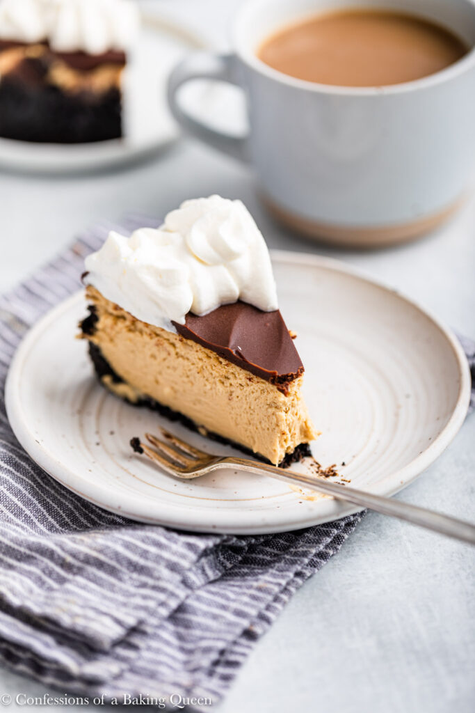slice of baked coffee cheesecake on a white plate with a fork sitting on a blue linen on a grey surface with a cup of coffee and cheesecake in the background 