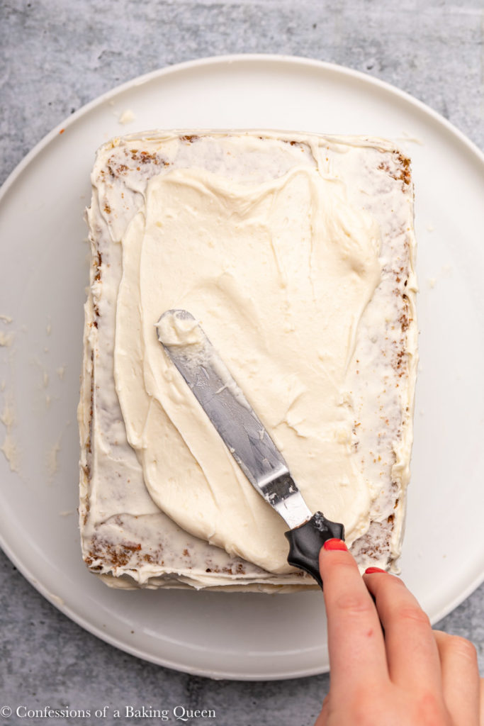 hand holding a spatula frosting a  cake with cream cheese frosting  on a grey background