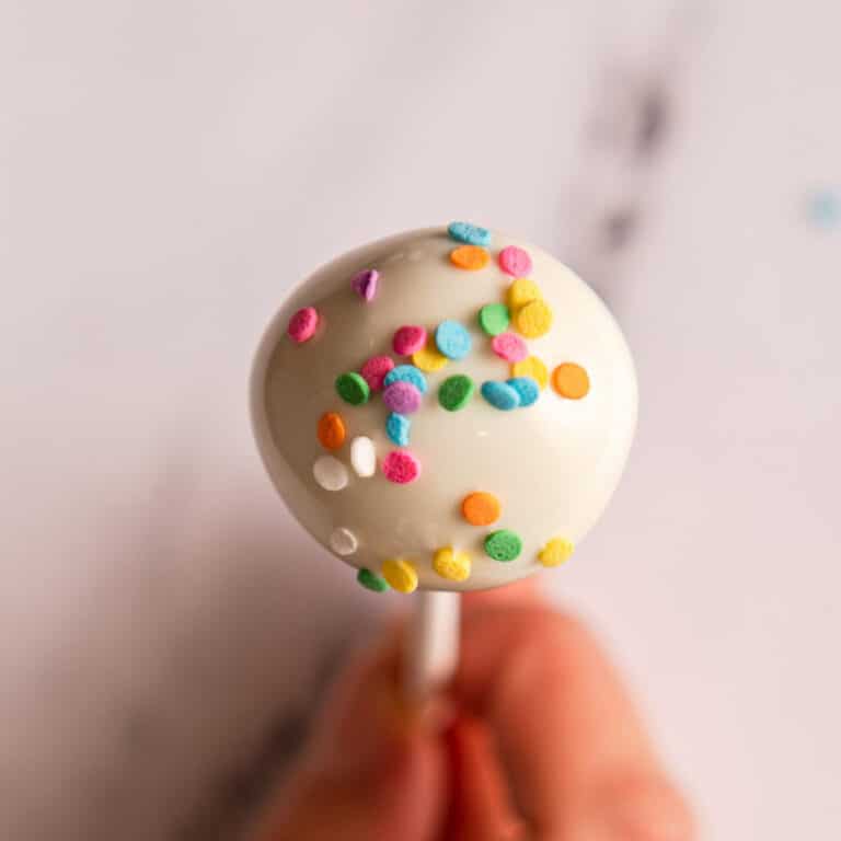 sprinkles added on top of white chocolate dipped cake pop on a white surface