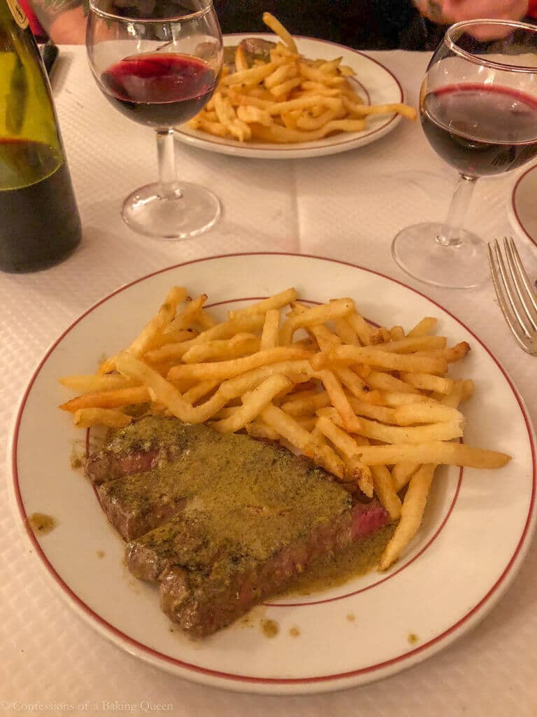 steak frites on aw hite plate next to a glass of wine on a dinner table