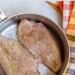 chicken breasts coated with salt and pepper in a silver saucepan on a grey surface with an orange linen