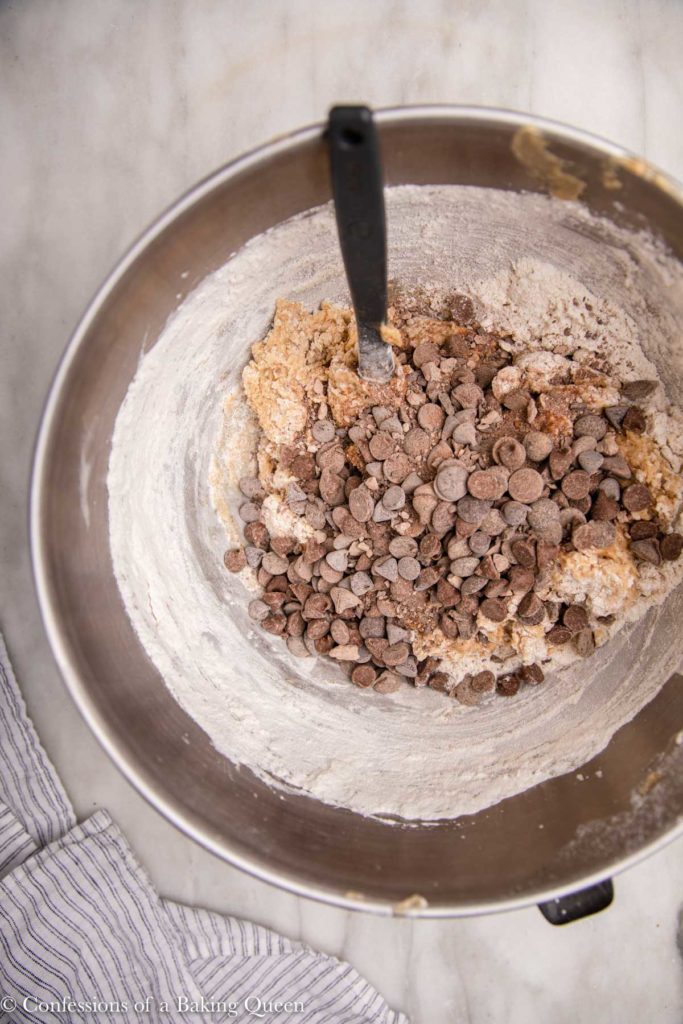 chocolate chips added to cookie batter in a metal mixing bowl on a white marble surface with a white stripped linen