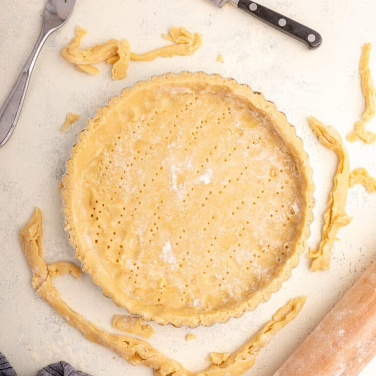 sweet shortcrust pastry in a tart tin before baking next to utensils on a white surface with a blue linen