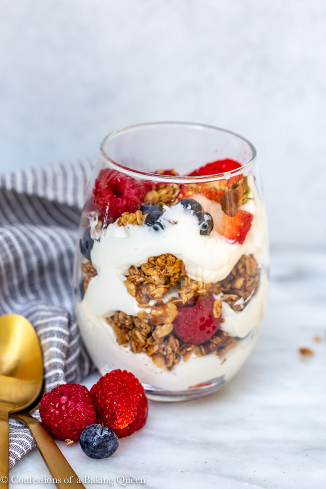 close up of yogurt parfait in a clear glass on a white marble background with gold spoons, a striped napkin and berries laying around 