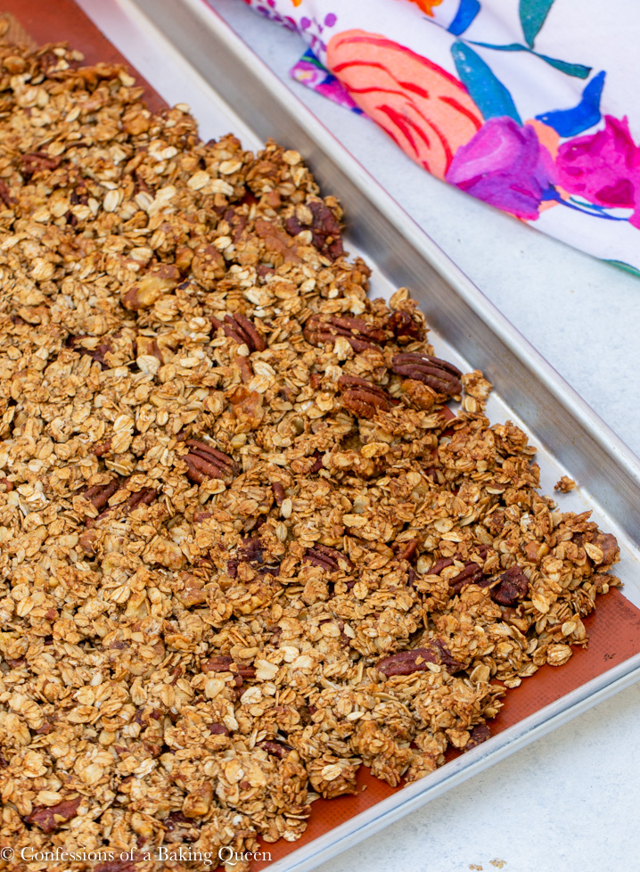baked banana nut granola cooling in metal pan with a colorful tea towel in top corner