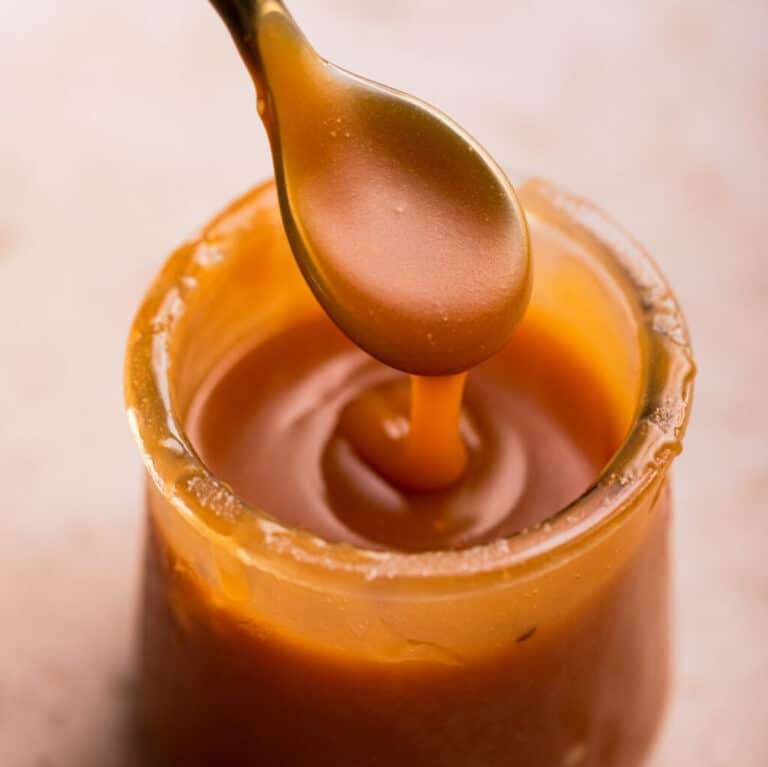 glass pot of salted caramel with a black spoon holding some dripping down sitting on a light brown surface